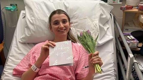 Kynleigh Parker is lying in a hospital bed smiling at the camera. She wears a pink hospital gown and holds a card in one hand and flowers in another. 