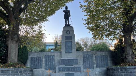 Pictured is the war memorial in St Peter Port. It is a stone structure with a statue at the top of it. It is surrounded by trees and grassland.

