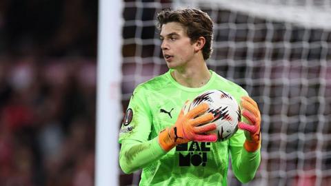 Melker Ellborg holding a ball in two hands close to his chest. He has dark hair and is wearing a lime green Malmo shirt and orange Puma goalkeeping gloves. His goal can be seen behind him.