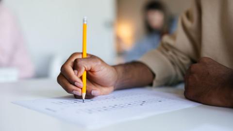 A close up of a male hand filling out a survey. They are holding a yellow pencil, leaning over the paper form. A person is in the background out of focus.