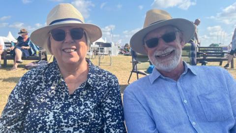 A man and a woman smiling at the camera. Both are wearing straw-type hats and sunglasses.