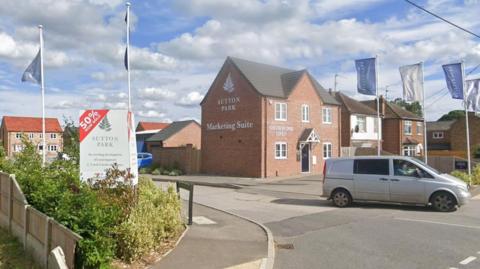 A road with a grey van waiting at a junction. Beside the road is a new red-brick building with a silver sign on it which reads "Sutton Park Marketing Suite" on one side and "Show Home Open" above a window on the facade. There are also blue and silver flags along the road and a sign which says "Sutton Park 50% sold". 