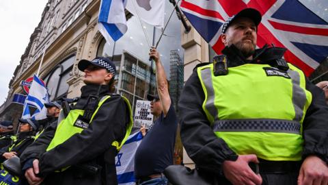 Police wearing yellow stand guard with a Union Jack flag visible in the background