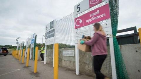 A woman carries a box as she crossed the car park of a recycling centre. A worker in a yellow high vis vest is seen entering the car park as well. The woman's figure is blurred. She is passing by a red sign that reads "Small appliances". A car with an open boot. It is a cloudy day.