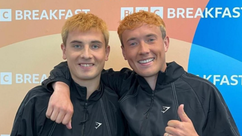 Cian Adams and brother Jordan are smiling. They are wearing black tracksuit tops and have dyed orange hair. They are standing in front of a BBC Breakfast logo.
