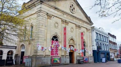 Corn Exchange Newbury is a tall, light stone building with carvings and topped by a triangular pediment. The ornate front entrance is flanked by a large arched window on either side.