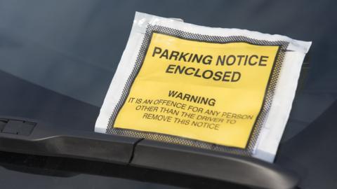 A parking enforcement notice in a yellow sleeve under a car's windscreen wiper