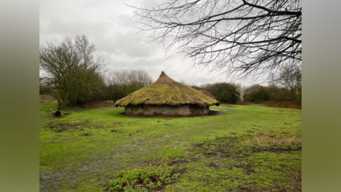 A roundhouse in a field surrounded by trees and shrubs.