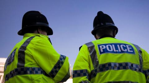 Two police officers with their backs to the camera in front of a blue sky. They both wear green high-vis jackets with "police" written on the back. Both wear black helmets.