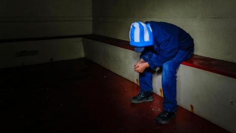 Stock photo of a boy wearing hood and blue jeans bent over with face hidden in a police cell