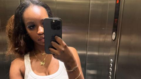 A young woman with curly dark hair looks directly into the camera in a close-up shot. She is wearing gold jewellery including a necklace and small hoop earrings. The purple BBC Radio WM logo is repeated across the backdrop behind her.

