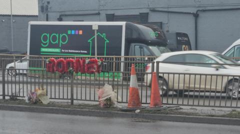 A metal crash barrier with a floral wreath attached to it. It's on a busy dual carriageway road with traffic and there are shop and office frontages in the background. The wreath is made of red flowers spelling out "Rodney". Two bouquets of flowers are also visible on the ground, and two traffic cones sit next to the flowers.