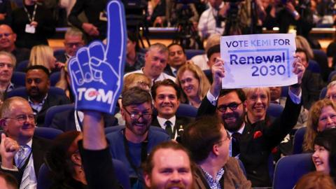 Bearded man in the audience at the Conservative Party conference waves Tom Tugendhat foam finger