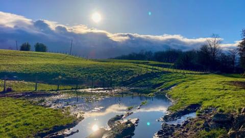 Bright sunshine above a layer of clouds in a blue sky with a field below with a big puddle