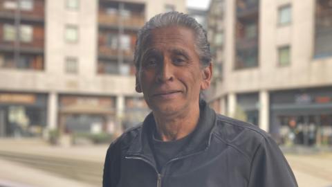 Michael Cunsamy has got grey slick-backed hair.. He is wearing a black zip jacket with a black top underneath. 