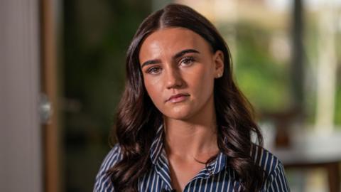 Sasha looking at the camera with a serious expression. She has long brown curly hair and wears a navy and white shirt. She is sat in her living room with the background blurred behind her.