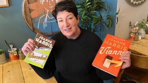 A woman with short dark hair sits in a cafe holding up two books which she has written.