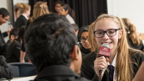 Female student with School Report microphone surrounded by other young people