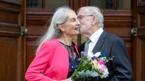 Elderly woman in a pink jacket and man in a suit kissing on the cheek