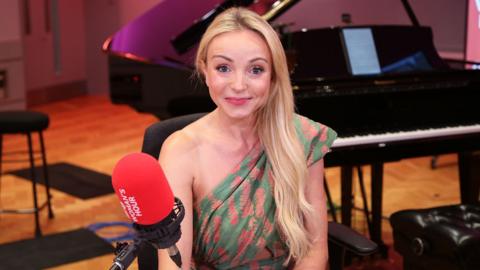 Helen George smiling behind a Woman's Hour-branded red microphone, with a piano visible in the background