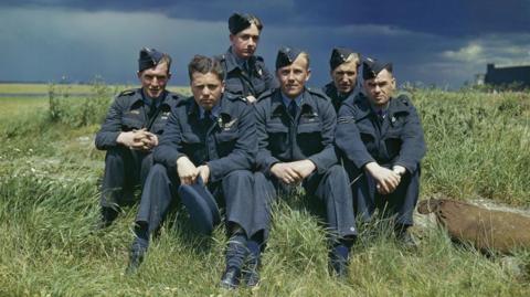 A colourised image of the 617 Squadron. They are sat in a field. Six men are in their blue RAF uniforms.