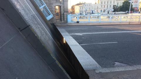 The east-side span of the Haven Bridge in the partially lifted position, showing thousands of wooden blocks that form the road surface, which will be replaced. There is a patterned steel plate on the edge of the level road surface of the bridge approach, blue painted lattice-work cast iron railings and a stone bridge operations house. In the distance are some Victorian and Edwardian buildings.