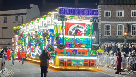 An illuminated carnival cart driving down a high street with crowds behind barriers on the pavement. The cart has Bourbon Street New Orleans in brightly-coloured lights on the front and more coloured lights all over it.