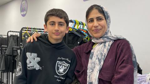 Hozan wearing a purple coat next to her son Younis in a black hoodie, in front of a row of uniform on rails