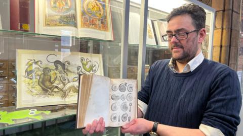 A middle-aged man with dark hair, glasses and a short beard holds a book open in front of a glass cabinet containing drawings of animals. The book itself is open on a page with drawings of fossils. The man is wearing a blue jumper over the top of a white shirt.