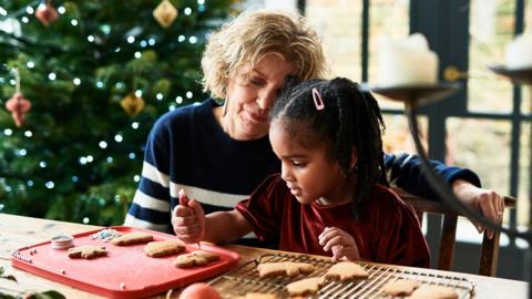 A grandmother helping her granddaughter ice gingerbread men. Behind them is a Christmas tree