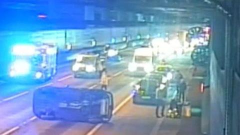 A car on its side in a tunnel on the A1. It is resting across a lane lengthways. Traffic is behing held behind it. On the right are people in high vis jackets. 