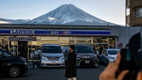 An image showing a photo being taken of a person standing in front of a Lawson shop, with Mount Fuji in the background