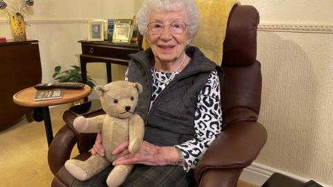Gabriele Keenaghan sitting in a chair with a teddy bear on her lap. It was the bear she brought with her from Vienna on the Kindertransport