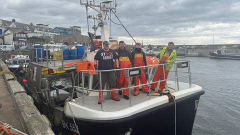 Four men are standing on a boat with Seahouses harbour behind them. They are all wearing waterproofs