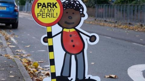 A sign which is a cartoon of a school child holding up a sign which reads PARK AT MY PERIL!