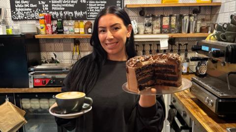 A woman with long black hair and an oversized black jumper is smiling into the camera whilst holding up a big chocolate cake which has been cut in half in one hand and in the other she is holding a black cup of latte in a saucer. Behind her is a coffee machine, a fridge, various drinks and teabags and a board with a menu on it written in white.