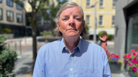Deputy Charles Parkinson - An older man with long grey hair, looking at the camera with a serious face. He is wearing a blue shirt with the top button undone.