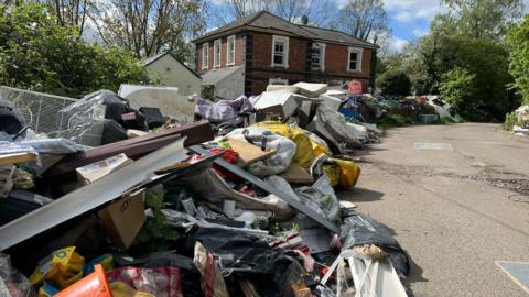 The old station, now with broken and boarded up windows. All along the road in front of it, and around the building, is flytipping - mattresses, white goods, blankets, and all kinds of rubbish. There is a red For Sale sign in front.