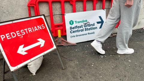 A sign saying Belfast Grand Central Station in navy letters on the ground beside an orange roadworks barrier and a red pedestrians sign. 