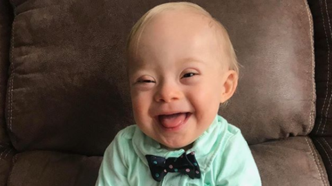 Lucas Warren, an 18-month-old baby with Down syndrome, smiles into the camera. He is wearing a light green shirt with a black spotted bowtie.