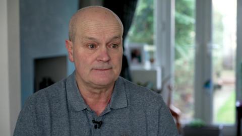 Vincent Ricci is pictured while being interviewed by the BBC. He is wearing a grey polo shirt while sitting in the living room of his home.