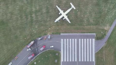 An aerial view of the white aircraft on the grass area of the runway it is facing horizontally compared to the direction of the runway itself. Multiple vehicles are seen parked on the runway.