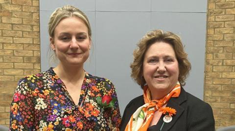 Two women, on the left Deputy Leader Lauren Townsend, and on the right Leader Jane Carr, stand side by side indoors in a formal setting, with a UK flag displayed behind them against a brick wall.