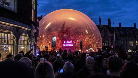 A monument in Durham Market Square is covered in a snowball like structure with snowflakes flying in it. A neon pink sign in front of the monument reads I LOVE DURHAM. A big crowd of people has surrounded the structure.