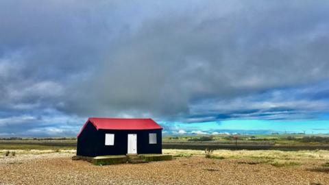 A black hut with a red roof sits on shingle with a cloudy blue/grey sky above