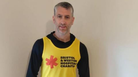 A man in his 40s with grey hair stands against a white wall wearing a black hoodie with a yellow Bristol and Weston Hospitals Charity vest top on.