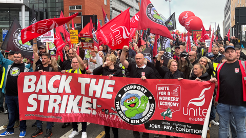 A crowd of people are holding red flags bearing the word Unite while the row at the front are holding a large banner bearing the words 'back the strike'