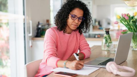 A young woman with black curly hair on a laptop at her kitchen, writing notes.