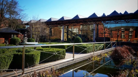 A view of the Streetlife museum building in Hull from across the landscaped gardens. There is a pond containing aquatic plants in the foreground which are lined with decorative hedges. The main building consists large glass panel walls and has a distinctive triangular roof.