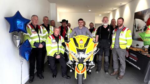 A group of Cornwall Blood Bike volunteers standing around an emergency motorbike